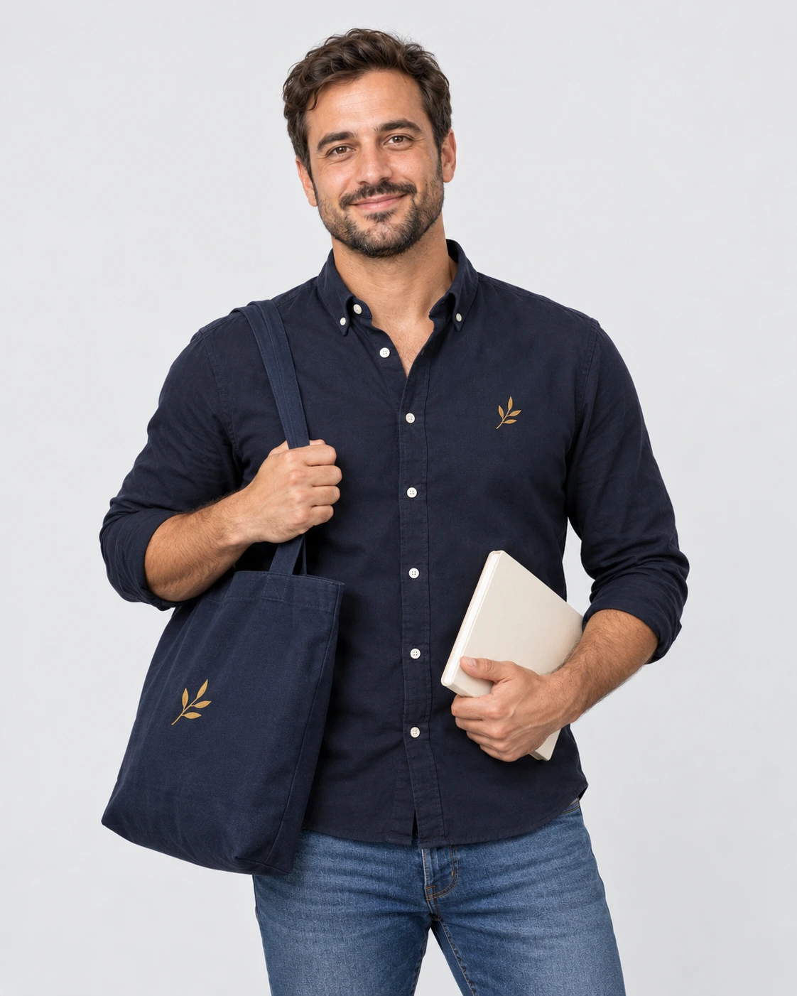 Man in a navy shirt holding a branded tote and notebook against a plain gray studio background