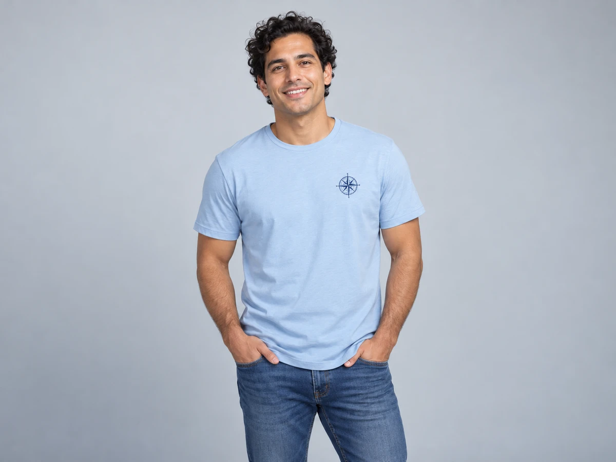 A 33-year-old Hispanic man with dark curls and clean shaven face wearing a pale blue T-shirt with a small crisp navy printed compass emblem on the left chest; casual confident pose.